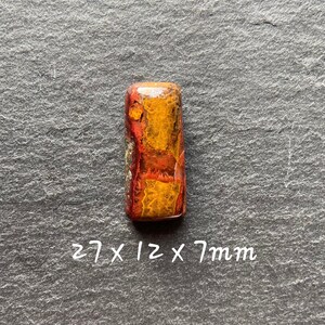 May include: A red, orange and yellow agate cabochon with a polished finish. The cabochon is rectangular and measures 27 x 12 x 7 mm.