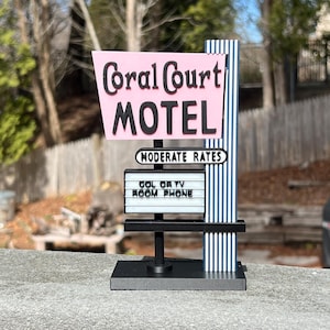 May include: A miniature Coral Court Motel sign with a pink and black sign. The sign includes the words "Coral Court Motel" and "Moderate Rates". The sign has a black base and a blue and white striped pole.