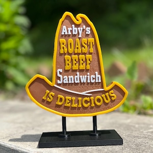 May include: A decorative sign in the shape of a cowboy hat, featuring the Arby's logo and the text "ROAST BEEF Sandwich IS DELICIOUS". The sign is brown, yellow, and white, mounted on a black stand.