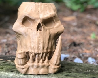 Puede incluir: Dos tazas de cerámica con forma de calavera, una con una gran sonrisa de dientes y la otra con un diseño de calavera más tradicional. Ambas son de color marrón claro.