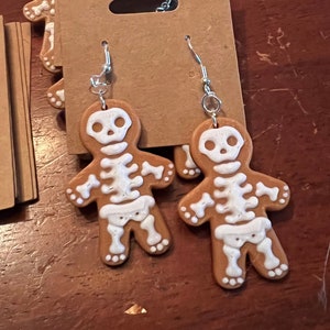 May include: A pair of skeleton gingerbread man earrings. The earrings are brown with white skeleton details and are hanging from silver hooks.