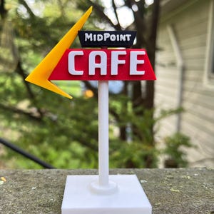 Puede incluir: Un letrero de cafetería de carretera en miniatura con una flecha amarilla, un letrero rojo con letras blancas que deletrean "CAFE" y la palabra "MIDPOINT" arriba. El letrero está montado sobre un poste y una base blancos, sobre un fondo exterior borroso.