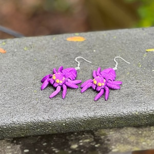 May include: A pair of purple spider earrings with yellow accents. The earrings are hanging from a silver hook.