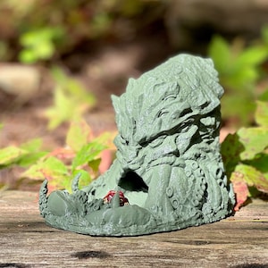Puede incluir: Una torre de dados impresa en 3D de color verde con forma de cabeza de monstruo con una abertura en la boca para tirar los dados. La torre está sobre una superficie de madera.