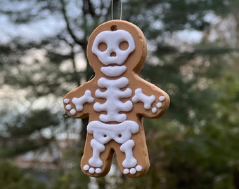 Puede incluir: Una galleta de jengibre con forma de esqueleto, con detalles de glaseado blanco. La galleta está colgada de una cuerda sobre un fondo borroso de árboles y una casa.