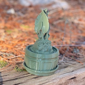 May include: A green 3D printed figurine of a small, leafy creature with a face, sitting in a pot with a decorative base.