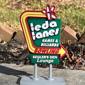 May include: A colorful sign for Ieda Lanes, a bowling alley and game room. The sign features a green background with white text and a yellow and red border. The sign also includes the text "Kegler's Den Lounge."
