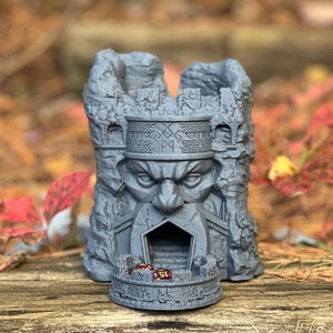 May include: A gray, fortress-shaped dice tower with a carved face and intricate details. The tower has a circular base and a slot for dice. The number "15" is visible on a die inside the tower. The background is blurred with fall foliage.