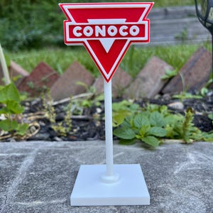 May include: A miniature Conoco sign featuring a red and white triangular emblem with the word CONOCO in white letters. The sign is mounted on a thin white vertical pole attached to a square white base, creating a freestanding display piece.