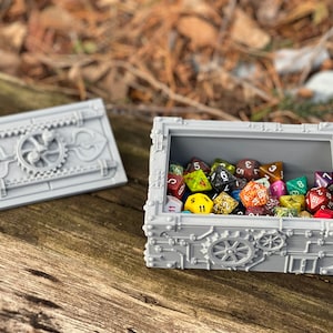 May include: A gray, metal, steampunk-style box with a hinged lid, open to reveal a collection of colorful dice. The box has intricate details, including gears and rivets.