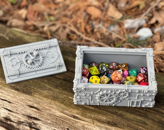 May include: A grey, metal, steampunk-style box with a hinged lid, open to reveal a collection of colourful dice. The box has intricate details, including gears and rivets.