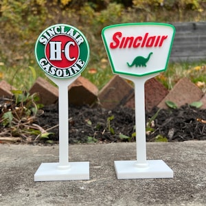 May include: Two miniature Sinclair gas station signs. One is round with "Sinclair" and "HC Gasoline" text, the other is a white sign with a green border, red "Sinclair" text, and a green dinosaur graphic.