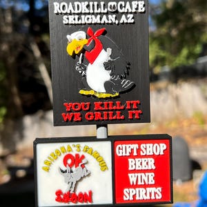 May include: A multi-layered sign for the Roadkill Cafe in Seligman, Arizona. The top sign features a cartoon vulture with the text "YOU KILL IT WE GRILL IT." The bottom sign advertises a gift shop, beer, wine, and spirits.