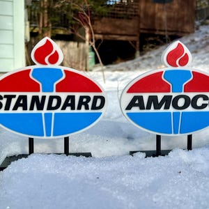 Puede incluir: Dos letreros de estilo vintage, uno con la palabra "STANDARD" y otro con "AMOCO". Cada uno presenta un diseño ovalado rojo y azul con una llama. Los letreros están montados sobre soportes negros y colocados en la nieve.