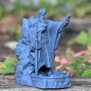 May include: A gray 3D printed miniature of a king standing in a rocky alcove. The king is wearing a crown and a long robe, and he is holding a sword in his left hand. His right hand is raised in a gesture of command. The miniature is designed to be used as a tabletop game piece.