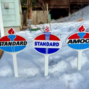 Puede incluir: Tres estacas de jardín de estilo vintage con los logotipos de las gasolineras Standard y Amoco. Cada estaca presenta un diseño ovalado rojo, blanco y azul con un emblema de antorcha y el nombre de la marca. Las estacas blancas están en la nieve.
