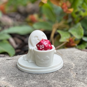 Puede incluir: Una figurita de fantasma blanco sosteniendo un dado rojo de 20 caras en un cubo de calabaza. La figurita está sobre una base blanca.