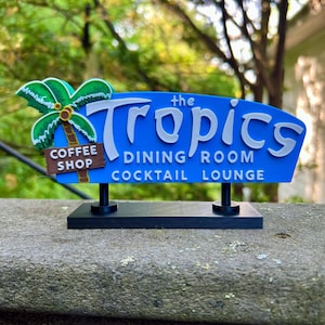 May include: A blue sign with the words "the Tropics" in white, with "DINING ROOM COCKTAIL LOUNGE" below. A palm tree graphic and "COFFEE SHOP" sign are on the left. The sign is mounted on a black base.