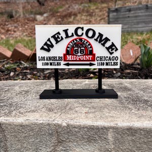 Puede incluir: Un letrero de bienvenida de la Ruta 66 con el texto "WELCOME" en negro, y "Los Ángeles 1139 millas" y "Chicago 1139 millas" debajo. El letrero presenta un emblema rojo y blanco que dice "Adrian, Texas, Route 66, Midpoint".