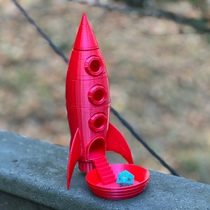 May include: A red 3D printed rocket ship with a blue and silver dice inside. The rocket has a small opening at the bottom with a ramp leading up to the opening.