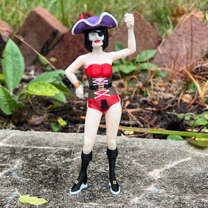 May include: A figurine of a female pirate, wearing a red and brown corset, red shorts, and black boots. She has a purple hat with a feather and an eye patch. The figure is standing on a gray surface with a green background.