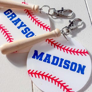 May include: Two baseball-themed keychains. Each keychain features a white circle with red stitching and a blue name. One says "GRAYSON" and the other says "MADISON". Each keychain has a small wooden baseball bat and a silver clasp.