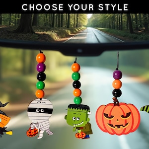 May include: Halloween-themed car charms hanging from a rearview mirror. Each charm features a different character: a witch, a mummy, Frankenstein, a jack-o'-lantern, and a black cat. The charms are strung with colorful beads and are set against a blurred background.