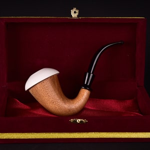May include: A wooden tobacco pipe with a white top and black stem rests inside a red velvet box with gold trim. The pipe has a curved design. The box is closed with a small gold crown.