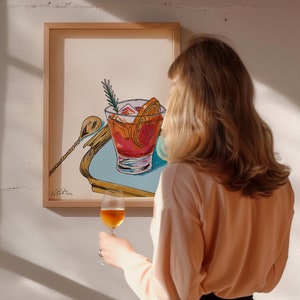 Puede incluir: Una ilustraci&oacute;n enmarcada de un c&oacute;ctel con una rodaja de naranja y una ramita de romero. La ilustraci&oacute;n es de estilo moderno con colores y l&iacute;neas atrevidas. La obra de arte est&aacute; colgada en una pared blanca.