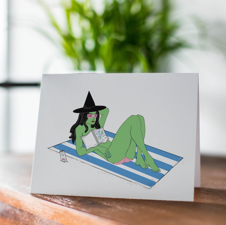 May include: A green witch wearing a black witch hat and pink sunglasses is lying on a blue and white striped beach towel. She is reading a book and has a bottle of sun cream next to her.