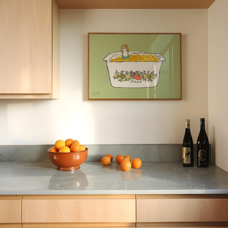 May include: A kitchen scene featuring a framed artwork with a woman in a tub of chips, a terracotta bowl filled with oranges, and two wine bottles. The artwork is framed in a light brown frame. The countertop is gray.