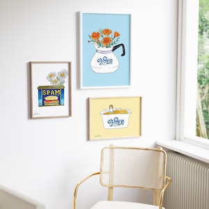 May include: Three framed prints with a blue and white floral theme. The top print features a blue and white pitcher with orange flowers. The middle print features a can of Spam with a daisy on top. The bottom print features a blue and white casserole dish with a yellow background.