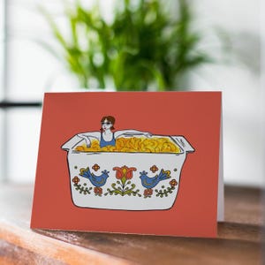 以下が含まれることがあります： 金色の食べ物で満たされたキャセロール皿の中でリラックスしている人のイラストが描かれたグリーティングカード。白い皿には青い鳥と花の模様があり、コーラル色の背景に設定されています。