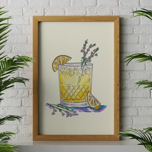 May include: A framed illustration of a yellow cocktail with a lemon wedge and a sprig of flowers. The drink is in a glass with a checkered pattern. The illustration is on a cream background.