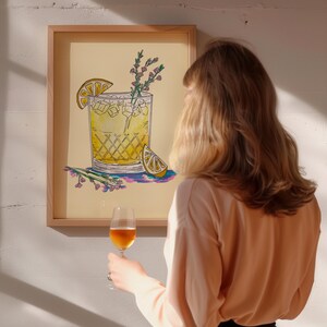 May include: A framed illustration of a yellow cocktail with a lemon wedge and sprigs of rosemary. The illustration is on a light yellow background and is framed in a light wood frame. A person is holding a glass of wine in front of the framed illustration.