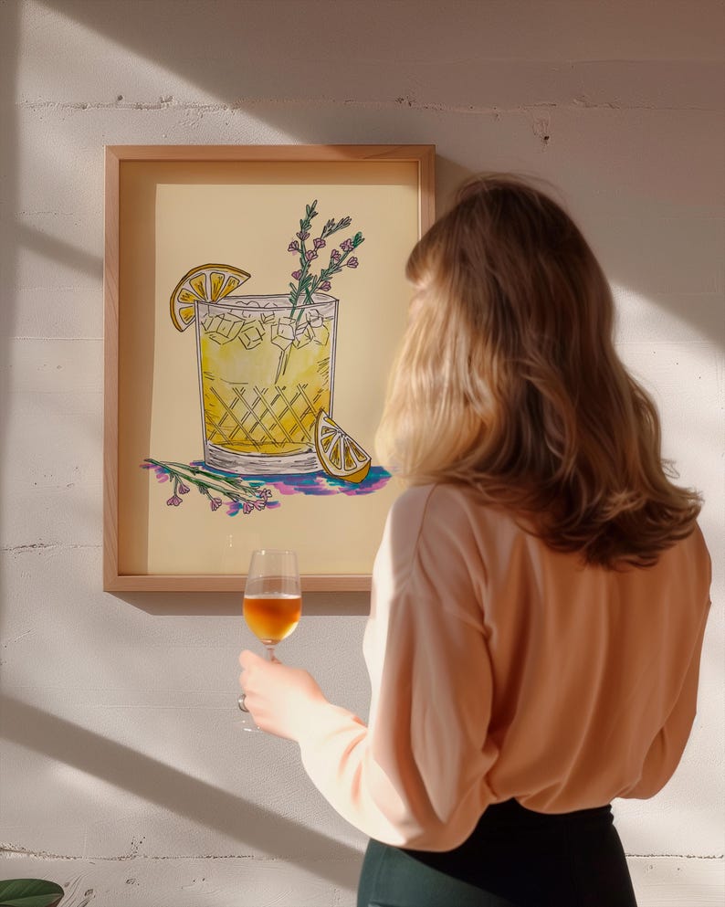 May include: A framed illustration of a yellow cocktail with a lemon wedge and sprigs of rosemary. The illustration is on a light yellow background and is framed in a light wood frame. A person is holding a glass of wine in front of the framed illustration.