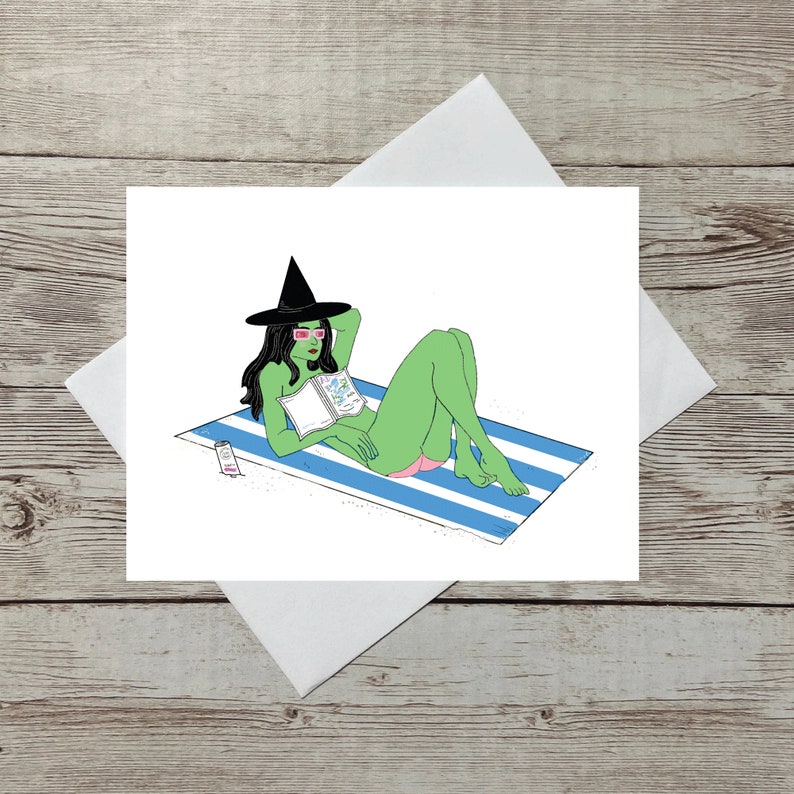 May include: A green witch wearing a black witch hat and pink sunglasses is lying on a blue and white striped beach towel. She is reading a book and has a bottle of sun cream next to her.