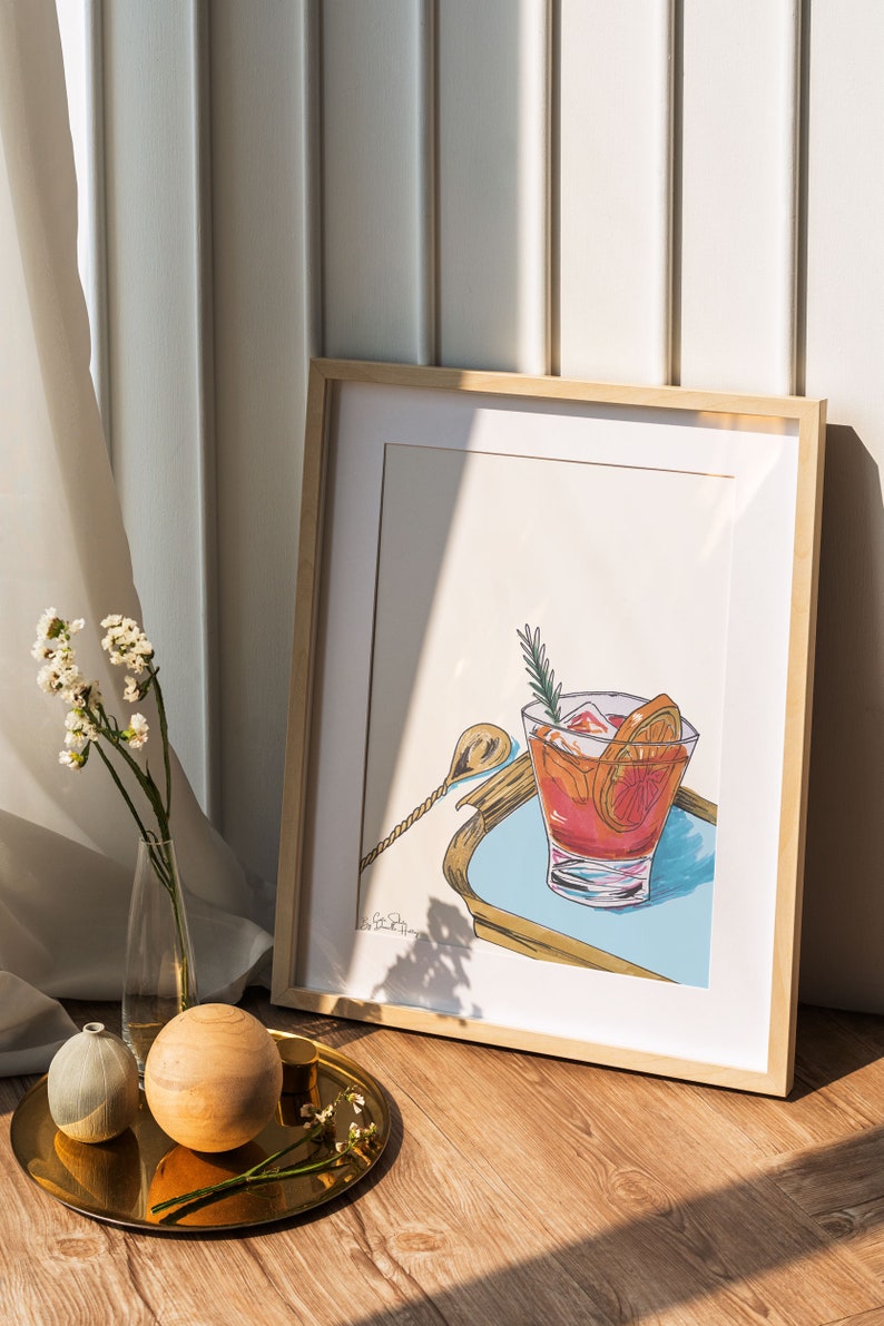 Puede incluir: Una ilustraci&oacute;n enmarcada de un c&oacute;ctel en un vaso con hielo, rodajas de naranja y una ramita de romero. El vaso est&aacute; sobre una bandeja azul y dorada con un agitador plateado.