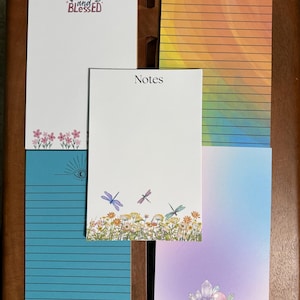May include: A selection of decorative notepads with diverse designs. One notepad features the text "Loved and Blessed" above a floral border. Other notepads have lined pages, watercolour backgrounds, and dragonfly or crystal illustrations.