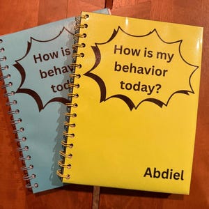 Puede incluir: Se muestran dos cuadernos con espiral, uno azul claro y otro amarillo. Los cuadernos presentan el texto "How is my behavior today?" en un diseño de bocadillo negro. El cuaderno amarillo está personalizado con el nombre "Abdiel".