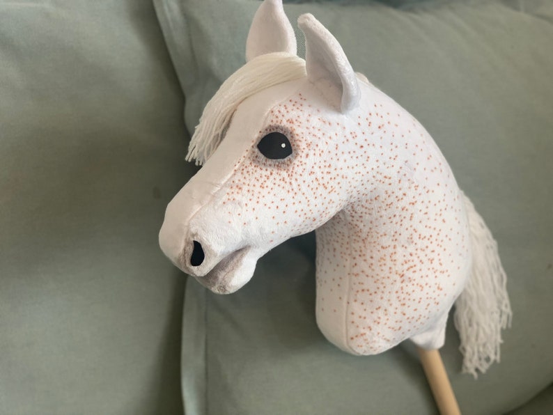 Puede incluir: Caballo de balanc&iacute;n de peluche blanco con manchas marrones y una crin y cola blancas. El caballo tiene ojos negros y un palo de madera para el cuerpo.