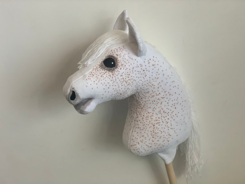 Puede incluir: Caballo de balanc&iacute;n blanco con manchas marrones. El caballo tiene una crin y una cola blancas. El caballo est&aacute; hecho de tela y tiene un palo de madera para el cuerpo.