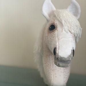 Puede incluir: Cabeza de caballo de peluche blanca con manchas marrones, una crin blanca y ojos negros. La cabeza del caballo est&aacute; unida a un palo de madera.