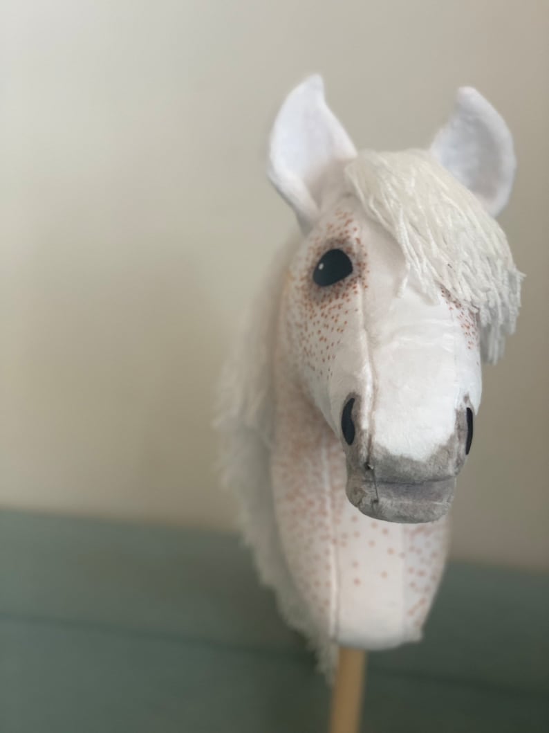 Puede incluir: Cabeza de caballo de peluche blanca con manchas marrones, una crin blanca y ojos negros. La cabeza del caballo est&aacute; unida a un palo de madera.