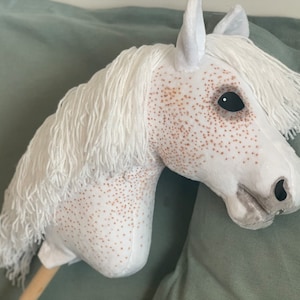 Puede incluir: Caballo de balanc&iacute;n de peluche blanco con manchas marrones y un ojo negro. El caballo tiene una larga melena y cola blancas.