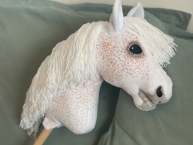 Puede incluir: Caballo de balanc&iacute;n de peluche blanco con manchas marrones y un ojo negro. El caballo tiene una larga melena y cola blancas.