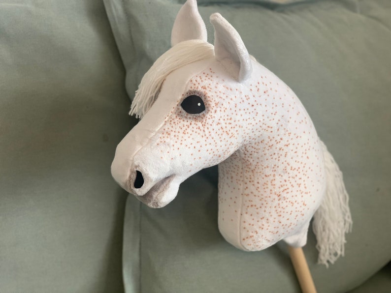 Puede incluir: Un caballo de balanc&iacute;n de peluche blanco con manchas marrones y ojos negros. El caballo tiene una larga melena y cola blancas.