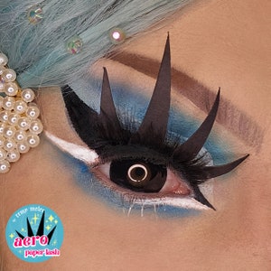 May include: Close-up of a person's eye with black dramatic eyelashes and a black circle contact lens. The eye is surrounded by blue and white eyeshadow. The text "aero paper lash" is visible in the lower left corner of the image.