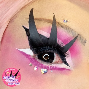 May include: Close-up of an eye with dramatic makeup. Black paper lashes and accents create a spiky effect. Pink and white eyeshadow, with rhinestones and a heart-shaped gem. The text "candy daggers paper lash" is visible.