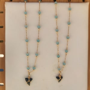 May include: Two gold necklaces with light blue beads and a black shark tooth pendant are displayed on a white surface. The necklaces are presented on a wooden display stand, with a beach background.